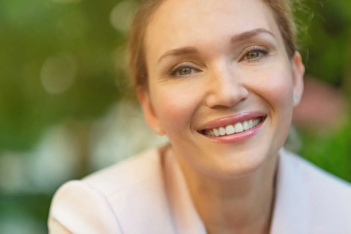 Close-up of a smiling woman with flawless teeth and glowing skin, outdoors with a blurred natural background, showcasing the confidence and beauty possible after receiving dental implants in Mokena IL