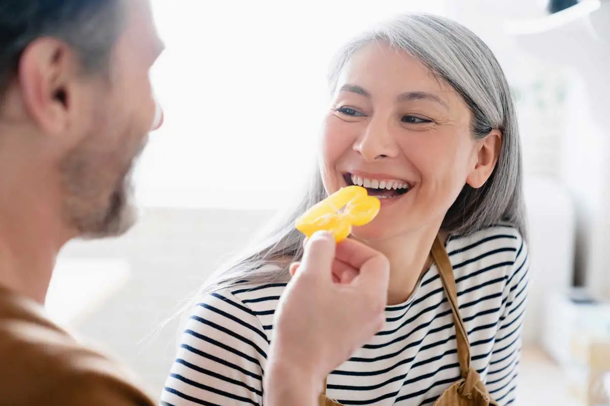 Smiling woman with long gray hair and striped shirt laughs joyfully as a man offers her a slice of yellow bell pepper in a sunlit kitchen, showcasing her healthy teeth, a vibrant moment highlighting the benefits of dental implants in Oswego IL.