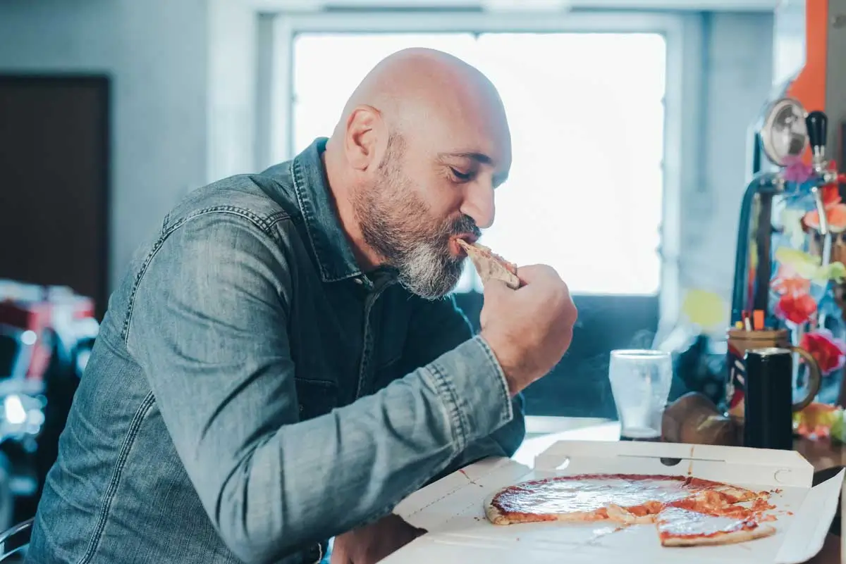 Middle-aged man with a bald head and gray beard enjoying a slice of pizza from a delivery box at a kitchen counter, confidently biting into the crust, showcasing the comfort and strength possible with dental implants in Mokena IL