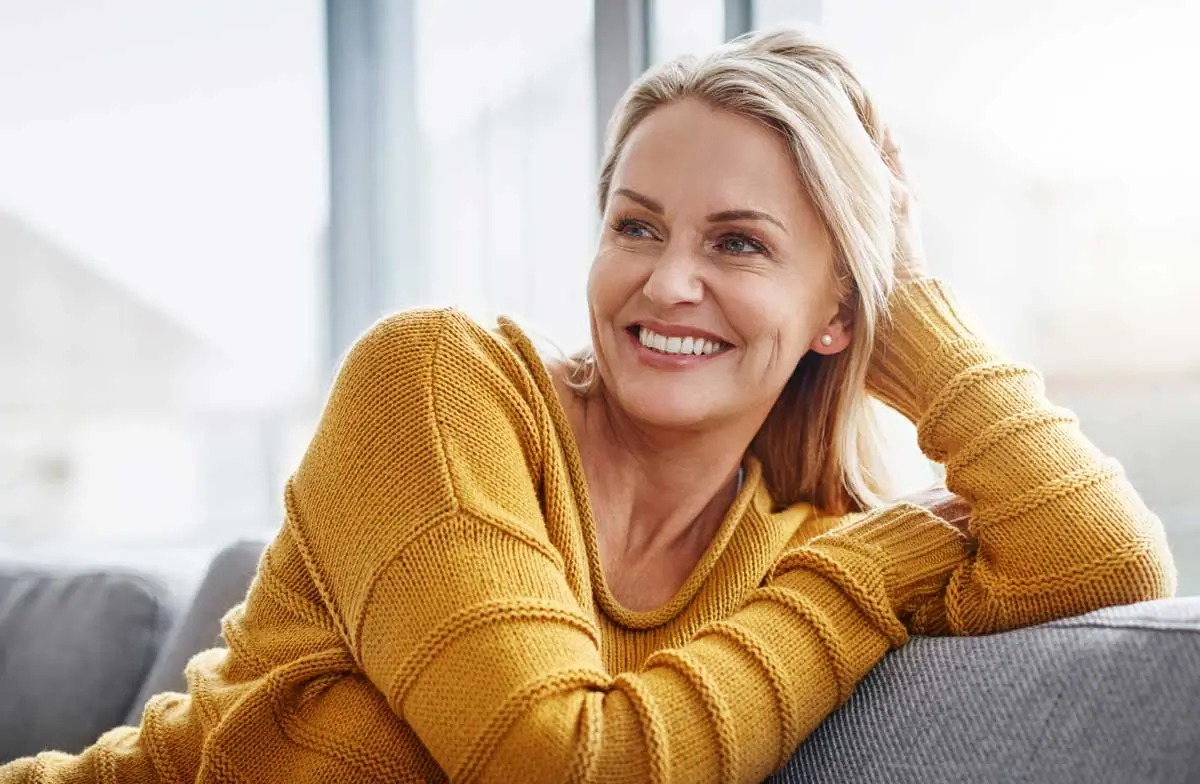 Middle-aged woman with radiant blonde hair and a cozy mustard sweater relaxes on a gray couch, smiling confidently with bright, healthy teeth, an ideal representation of the natural look and comfort offered by dental implants in Oswego IL.