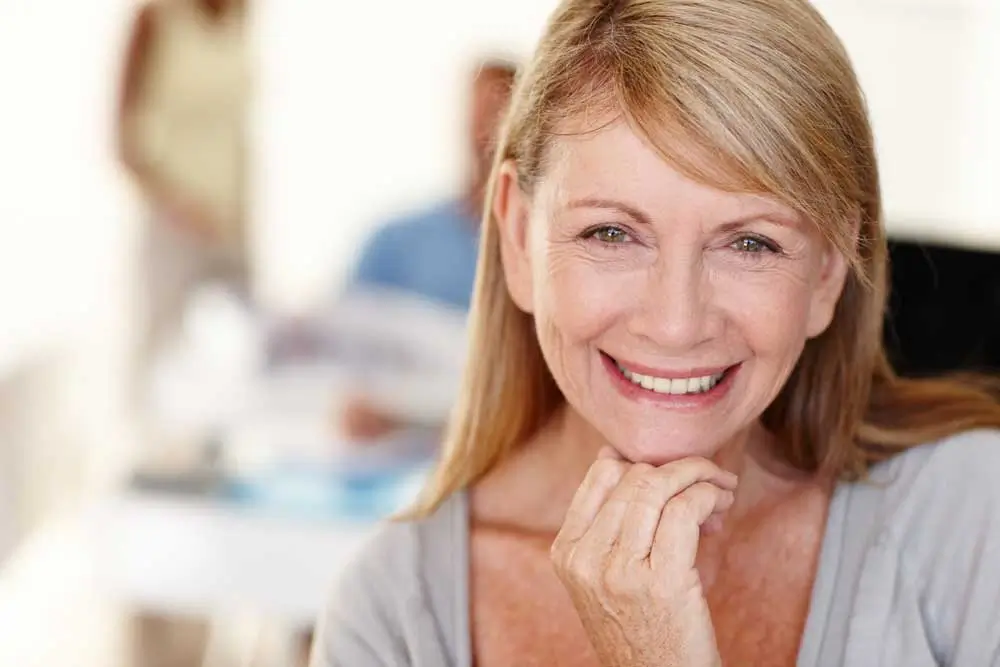 Confident, smiling middle-aged woman with straight blonde hair and clear skin, resting her chin gently on her hand in a well-lit office setting—her radiant smile highlights the natural-looking results of dental implants, perfect for promoting Dental Implants Shorewood IL