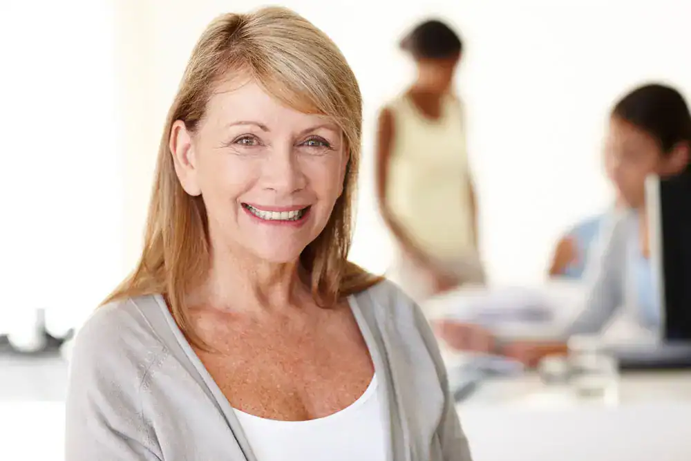 Smiling older woman with straight blonde hair wearing a light cardigan in a softly lit office environment, confidently showing her restored teeth—an example of natural-looking results from dental implants Westmont residents trust for a rejuvenated smile.