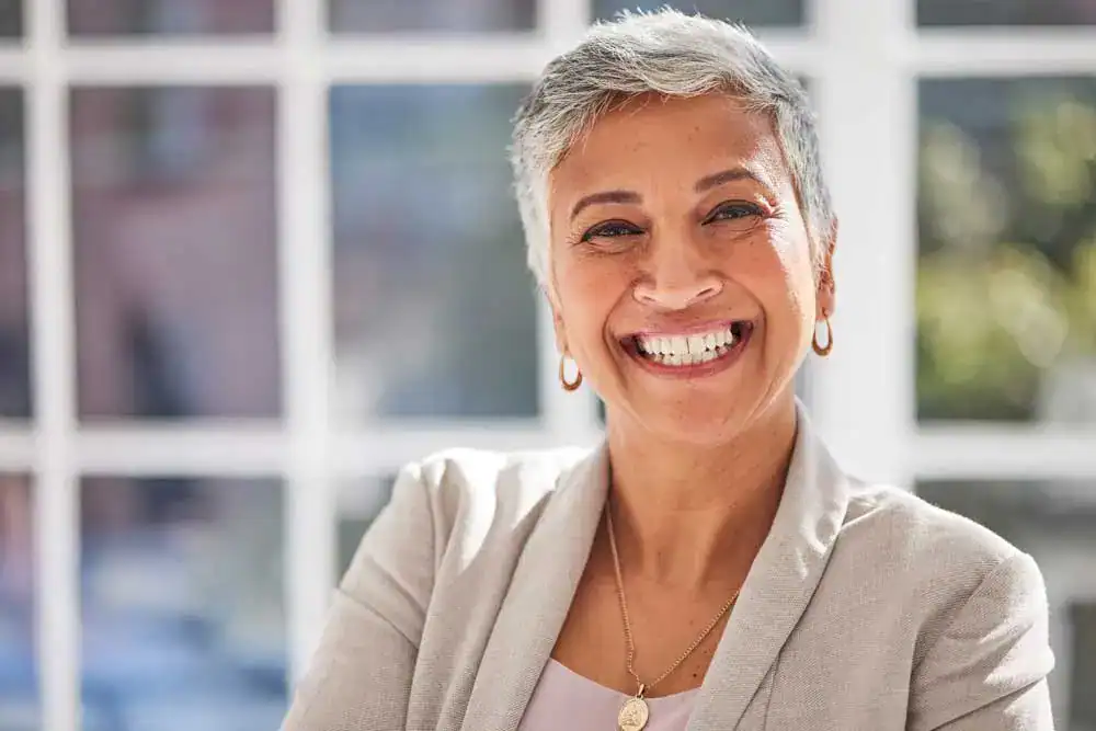 Mature woman with short gray hair and a radiant smile standing confidently in front of a bright window, showcasing the natural results of dental implants Tinley Park IL patients trust for restoring both function and aesthetics.