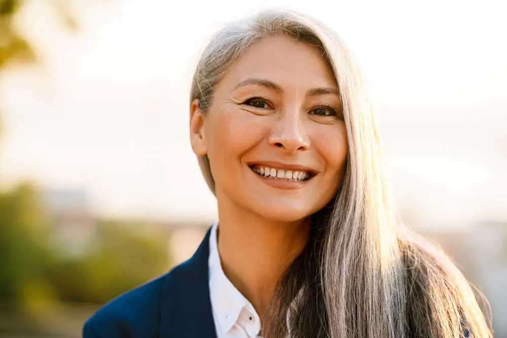Elegant middle-aged woman with long silver hair and a natural, confident smile, dressed in a navy blazer and white blouse, standing outdoors in soft golden light—her bright and healthy teeth reflect the lasting benefits of Dental Implants Shorewood IL