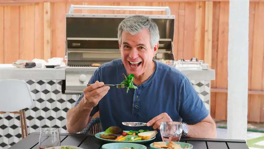 Cheerful middle-aged man with salt-and-pepper hair enjoying a fresh meal outdoors, confidently biting into leafy greens—his bright smile and ease while eating reflect the comfort and strength provided by dental implants in Darien IL.