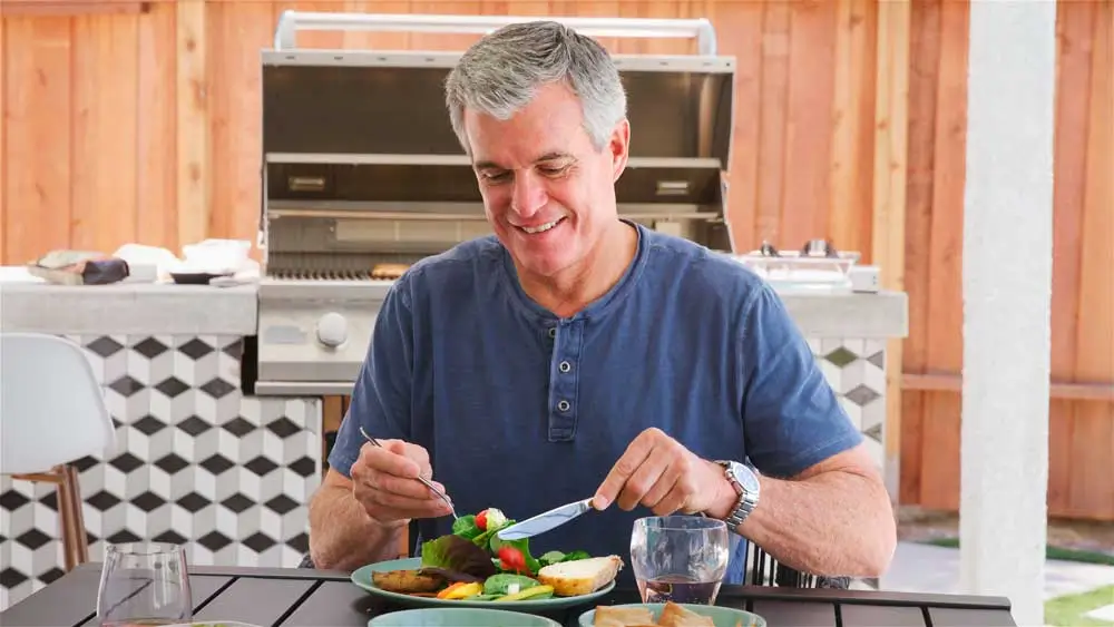 Smiling older man with short gray hair enjoying a fresh, colorful meal on a patio beside a stainless steel grill—his confident expression and ability to eat comfortably outdoors highlights the life-changing benefits of Dental Implants Crest Hill IL